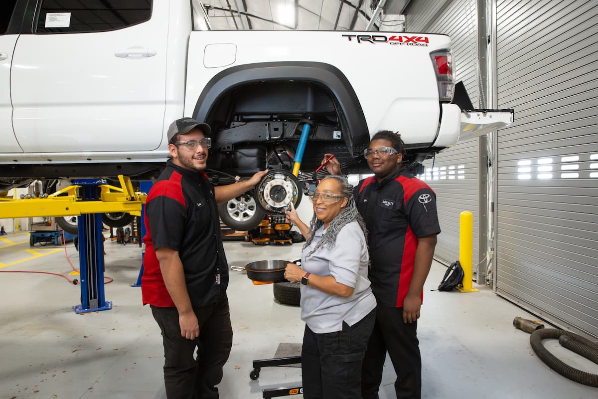 two male students learn from a black female instructor while working on a Toyota pickup truck